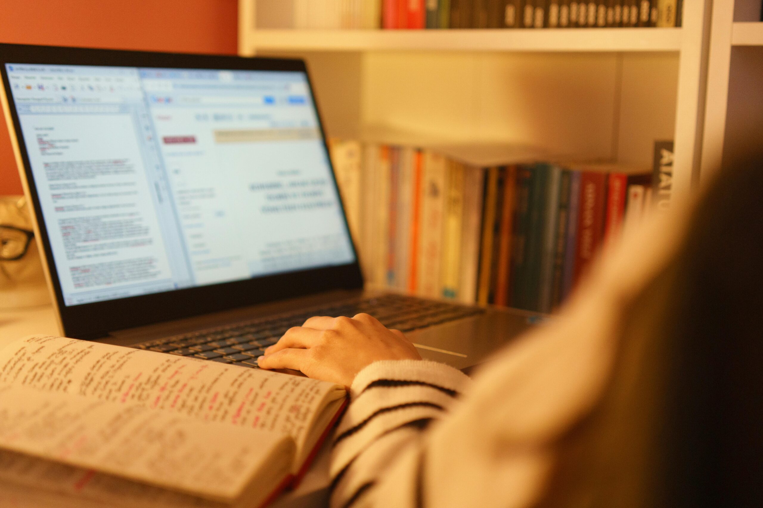 pexels-photo-31666035-31666035 A person studying online with a laptop and open book in a cozy home setting, surrounded by shelves of books.